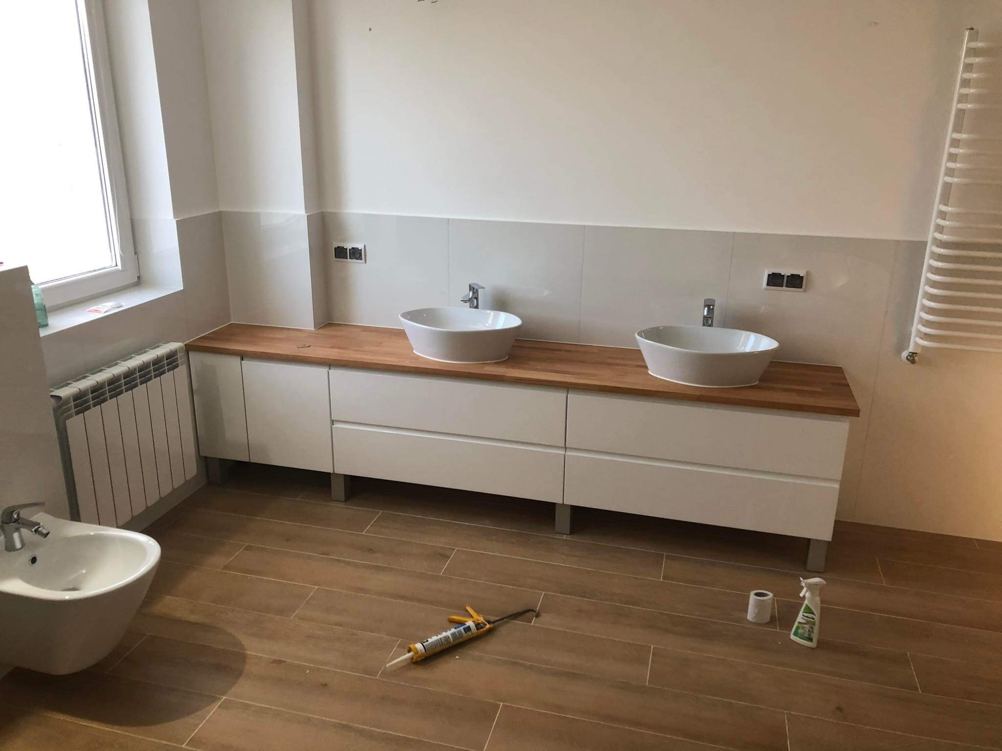 Nowoczesna łazienka z dwoma umywalkami nablatowymi, drewnianym blatem i brązowymi płytkami podłogowymi. Widoczny bidet i grzejnik drabinkowy.