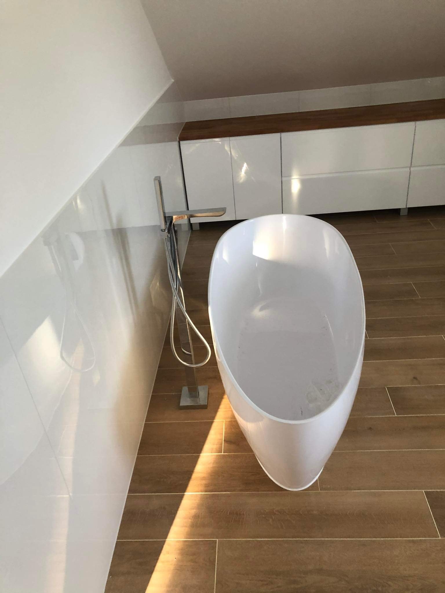 Nowoczesna, wolnostojąca wanna w łazience na poddaszu z drewnopodobną podłogą i białymi płytkami ściennymi. Bateria podłogowa. Białe szafki w tle.