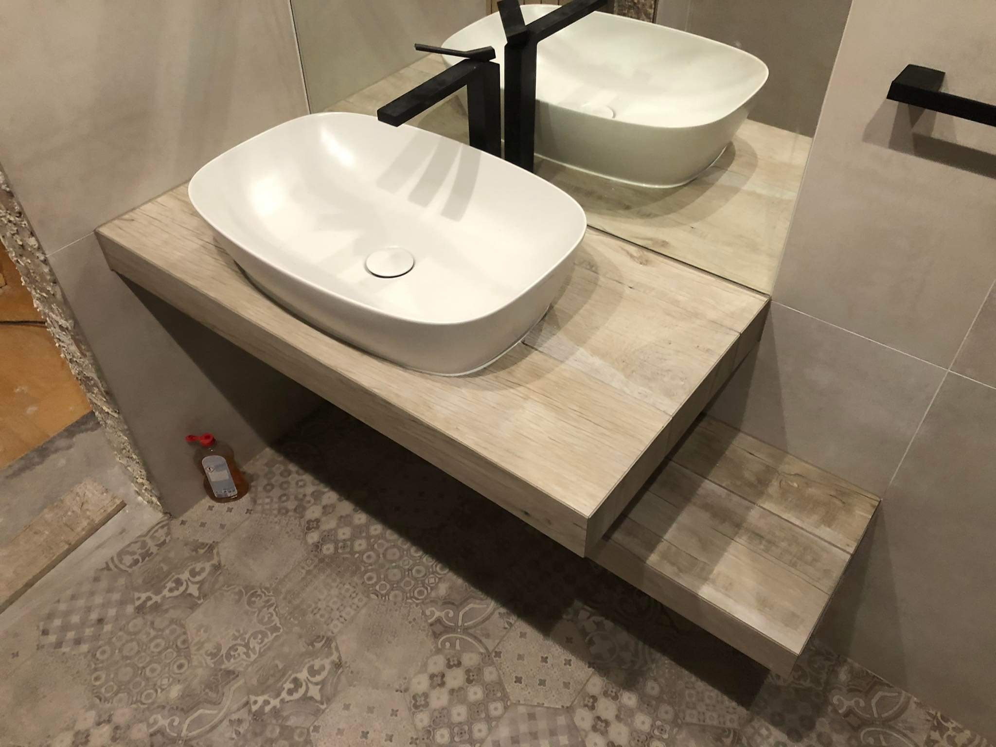 Nowoczesna łazienka z białą umywalką nablatową i czarną armaturą na drewnianym blacie. W tle lustro i płytki w odcieniach beżu. Na podłodze wzorzysta terakota.