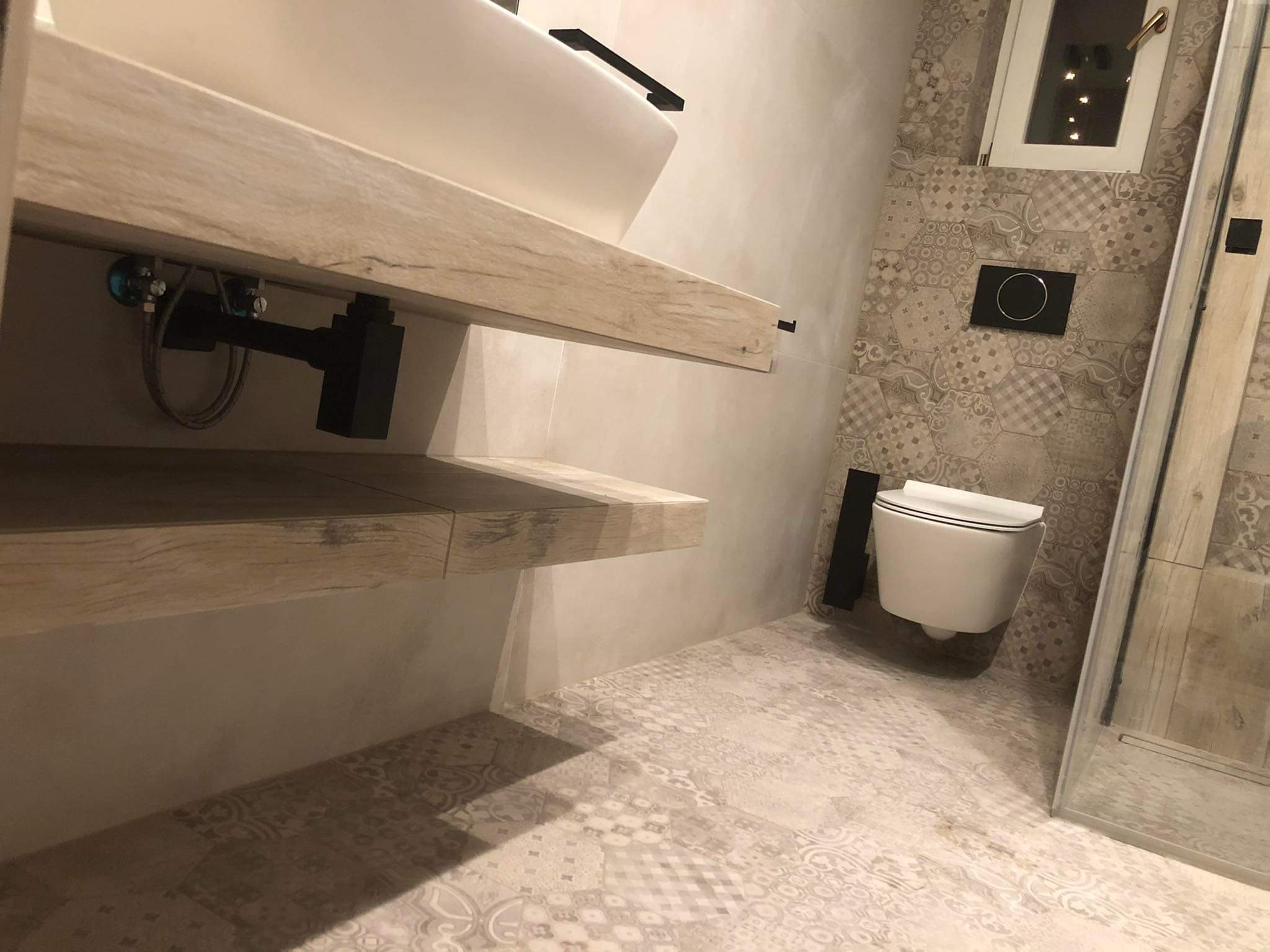 Nowoczesna łazienka z drewnianymi półkami pod umywalką, wzorzystą glazurą na ścianach i podłodze. Uwagę zwraca podwieszana toaleta i minimalistyczna armatura.