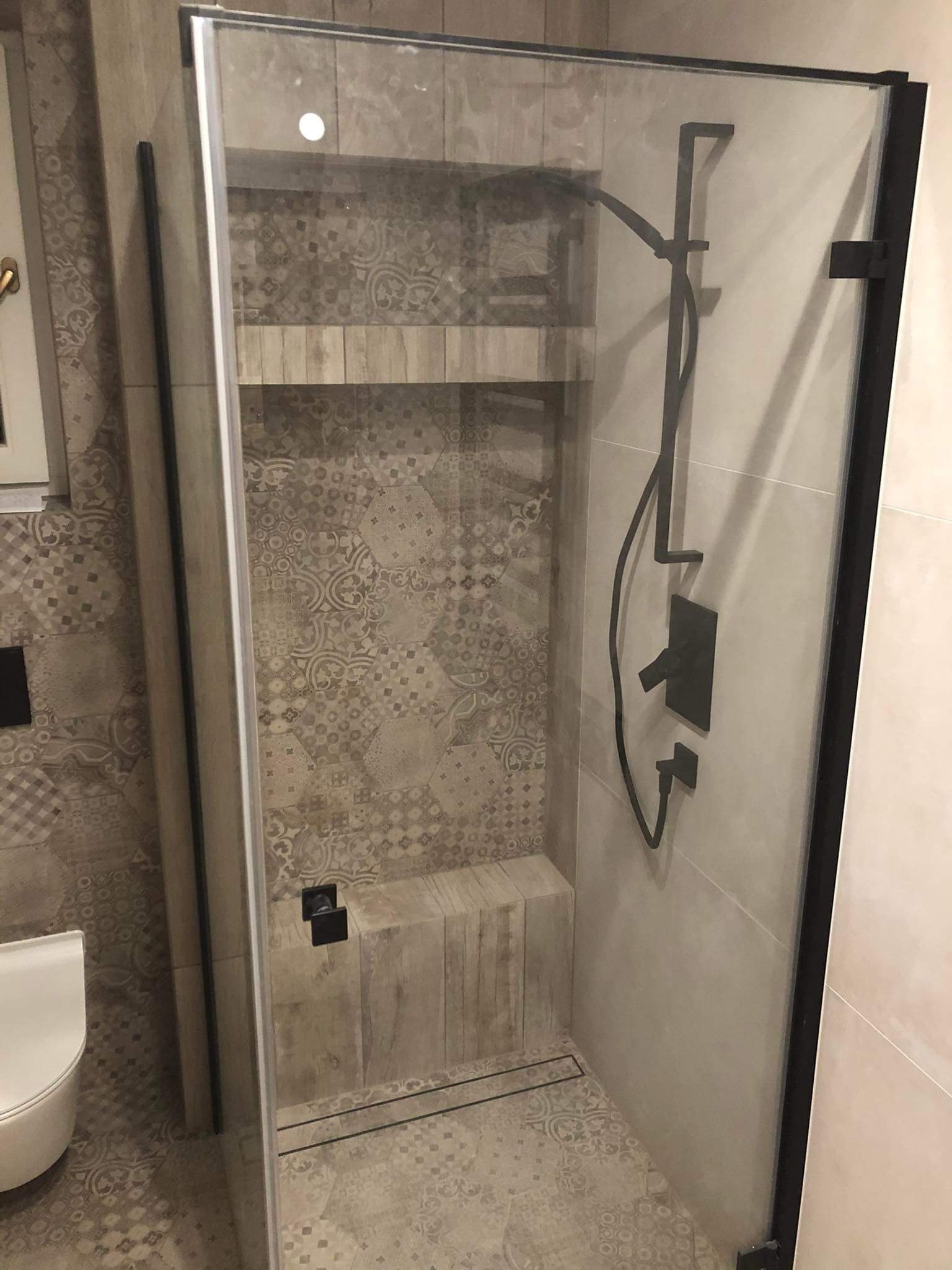 Kabina prysznicowa z czarną armaturą i wzorzystymi płytkami w beżach. Widoczna półka i siedzisko wewnątrz kabiny. Nowoczesny design łazienki po remoncie.