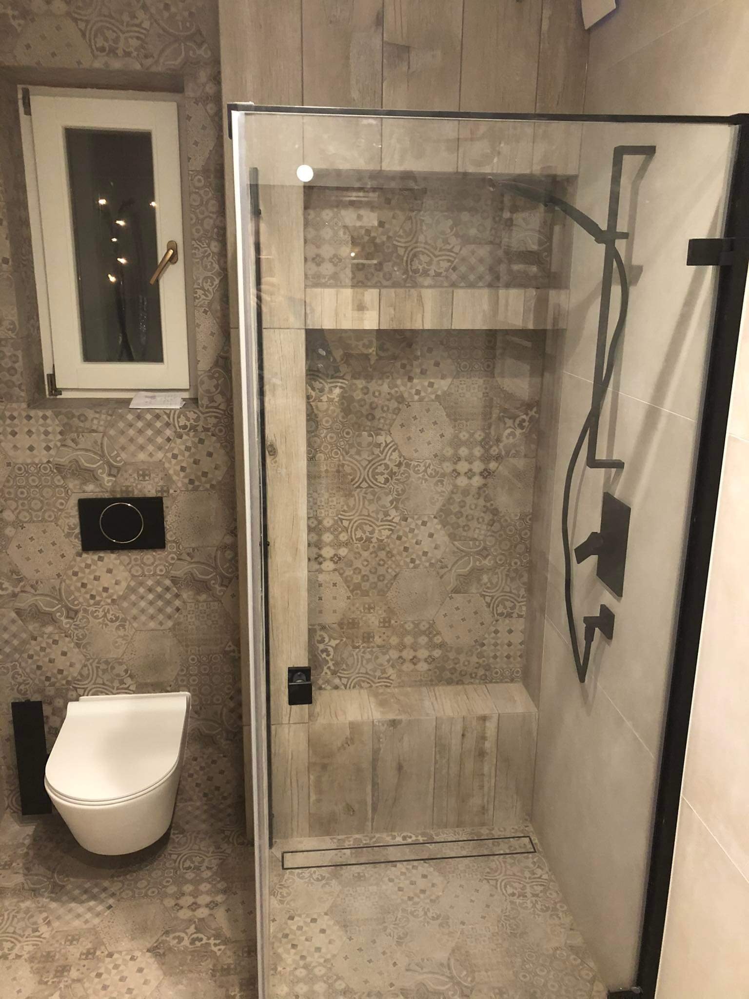 Nowoczesna łazienka z kabiną prysznicową typu walk-in, wykończona płytkami w odcieniach beżu i brązu z geometrycznym wzorem. Widoczna toaleta podwieszana i okno.