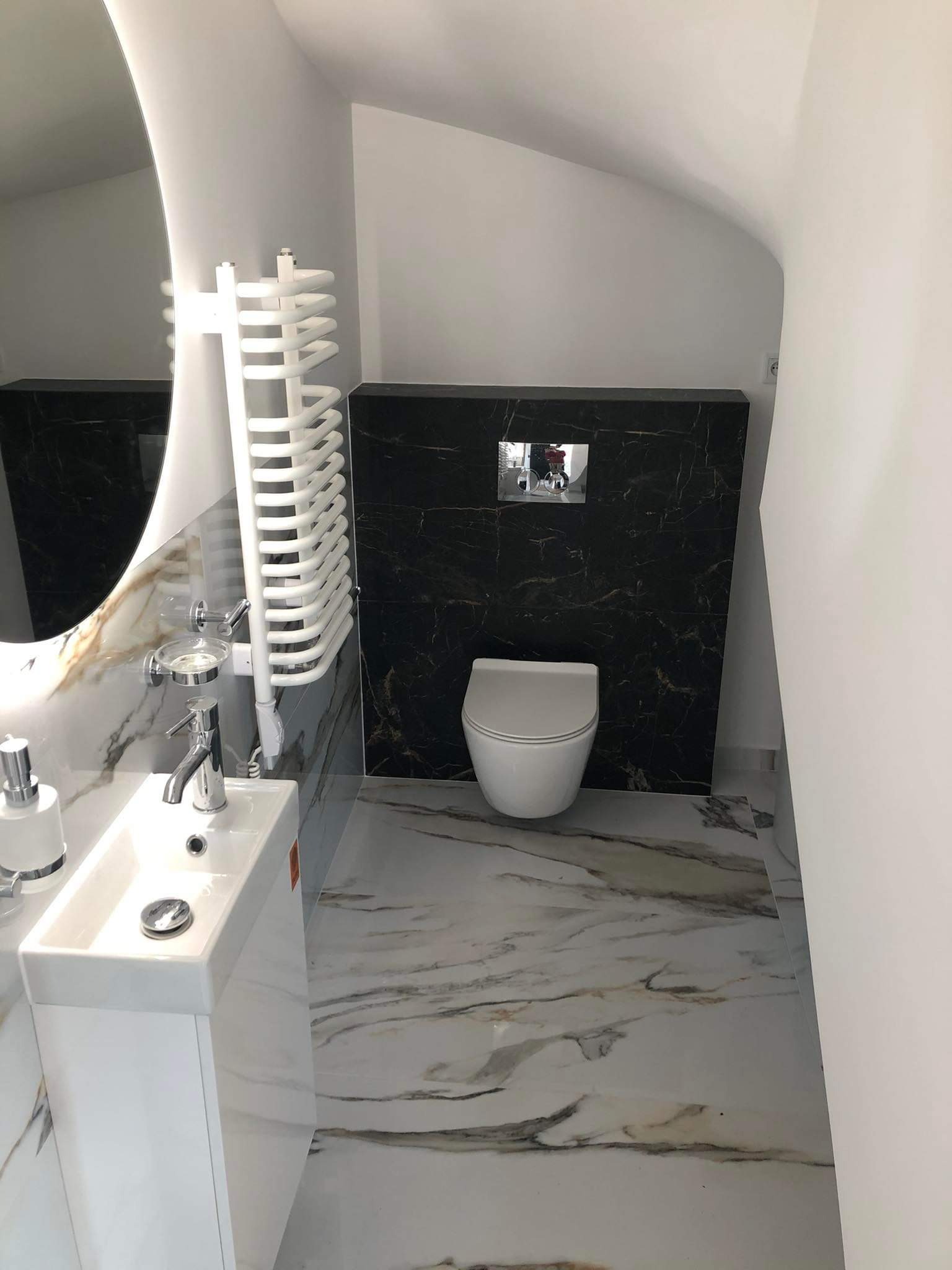 Nowoczesna łazienka w odcieniach bieli i marmuru z czarną ścianą akcentującą toaletę. Umywalka z szafką, owalne lustro i grzejnik drabinkowy.