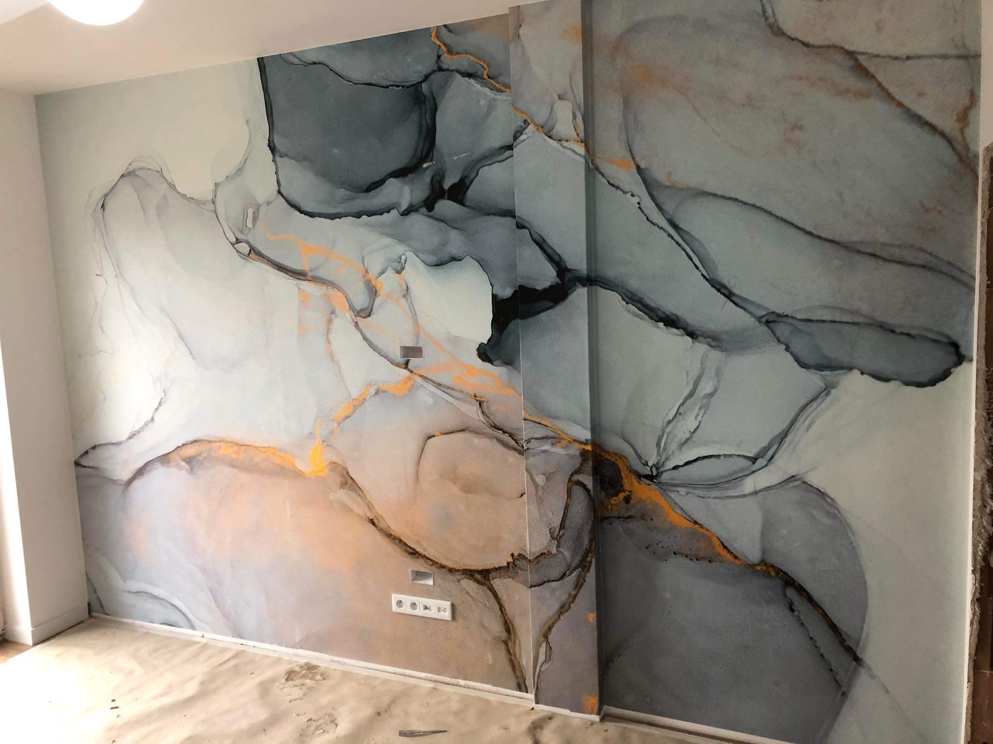 Ściana z unikalnym wzorem imitującym marmur w odcieniach szarości i złota, z widocznymi gniazdkami elektrycznymi i zabezpieczoną podłogą. Dekoracyjne wykończenie.