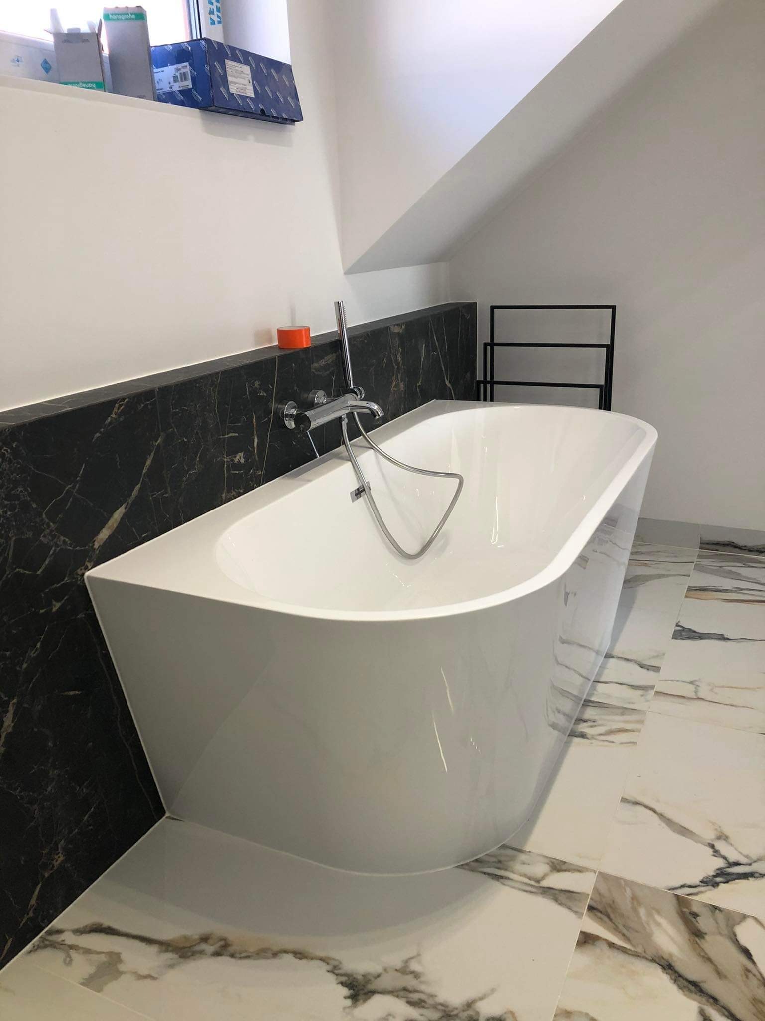 Biała wanna wolnostojąca z chromowaną armaturą na tle ściany z czarnego marmuru i podłogi z białych płytek imitujących marmur w łazience na poddaszu.