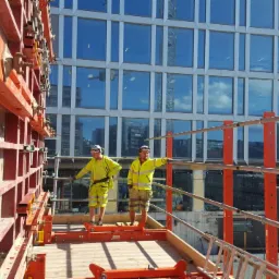 Dwóch pracowników w żółtych ubraniach roboczych stoi na platformie rusztowania na wysokości, z widoczną szalunkową konstrukcją i budynkiem w tle.