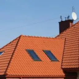 Czerwony dach z dachówki ceramicznej z dwoma oknami połaciowymi, komin z antenami na tle błękitnego nieba.