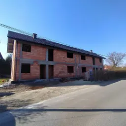 Stan surowy zamknięty bliźniaka w Myślenicach z cegły ceramicznej, widoczne okna i drzwi bez stolarki, dach pokryty dachówką, teren wokół budynku przygotowany do dalszych prac.