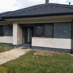 Dom jednorodzinny z ciemną dachówką karpiówką, elewacją w odcieniach szarości i beżu, widok z przodu, trawnik z liśćmi.