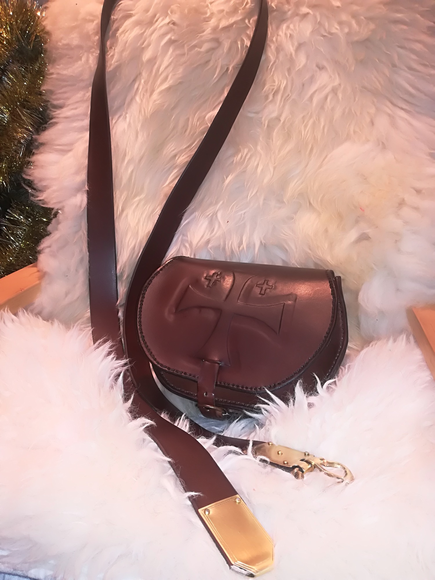 Brązowa skórzana torba z wytłoczonym krzyżem na klapie, z długim paskiem i ozdobnym zapięciem, leżąca na białym, puszystym futrze.