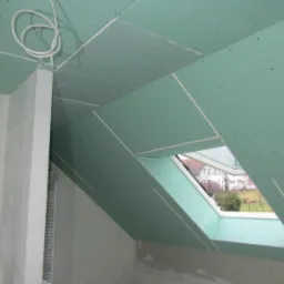 Montaż zielonych płyt karton-gips na poddaszu z oknem dachowym, widoczne łączenia i wkręty, instalacja elektryczna na suficie.