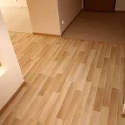 Ułożona podłoga z paneli imitujących drewno, przechodząca w płytki ceramiczne w sąsiednim pomieszczeniu, widok z perspektywy wejścia.