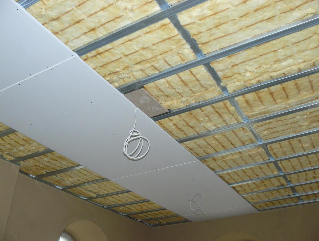 Montaż sufitu podwieszanego z płyt kartonowo-gipsowych, widoczna konstrukcja metalowa, izolacja z wełny mineralnej oraz częściowo zamontowane płyty, z wiszącym przewodem elektrycznym.