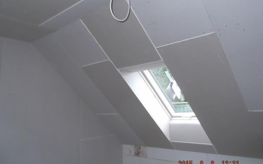 Poddasze z oknem dachowym w trakcie montażu płyt kartonowo-gipsowych, widoczne łączenia i surowe wykończenie.
