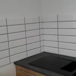 Białe, prostokątne płytki ułożone w rzędach z czarną fugą, czarny zlew i bateria kuchenna ze stali szczotkowanej, fragment okna w tle.