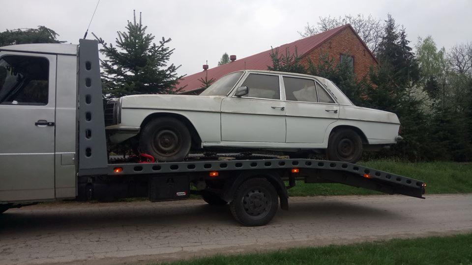 Biały, starszy model Mercedesa przewożony na platformie lawety w wiejskiej scenerii, z widocznym ceglanym domem w tle.