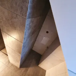 Ukośne ujęcie wykończonej łazienki z brązowymi i beżowymi płytkami, widoczny fragment toalety i klapa rewizyjna w suficie.