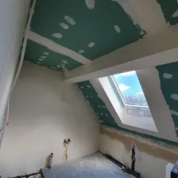 Poddasze w trakcie remontu: zielone płyty karton-gips z widocznymi śladami po wkrętach na skośnym suficie, okno dachowe z widokiem na niebieskie niebo, fragmenty starego muru, instalacje...