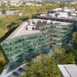 Nowoczesny, przeszklony budynek biurowy o trójkątnym kształcie, widok z góry, z widocznymi instalacjami na dachu, otoczony zielenią i torami kolejowymi.