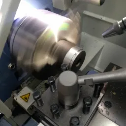 Metalowy element obrabiany na tokarce, widoczne wióry i narzędzie skrawające w trakcie pracy.