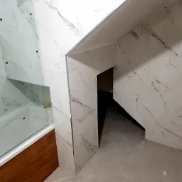 Wykończona łazienka z wanną za szklanym parawanem, marmurowe płytki na ścianach i szara podłoga, widoczny fragment wnęki pod schodami.