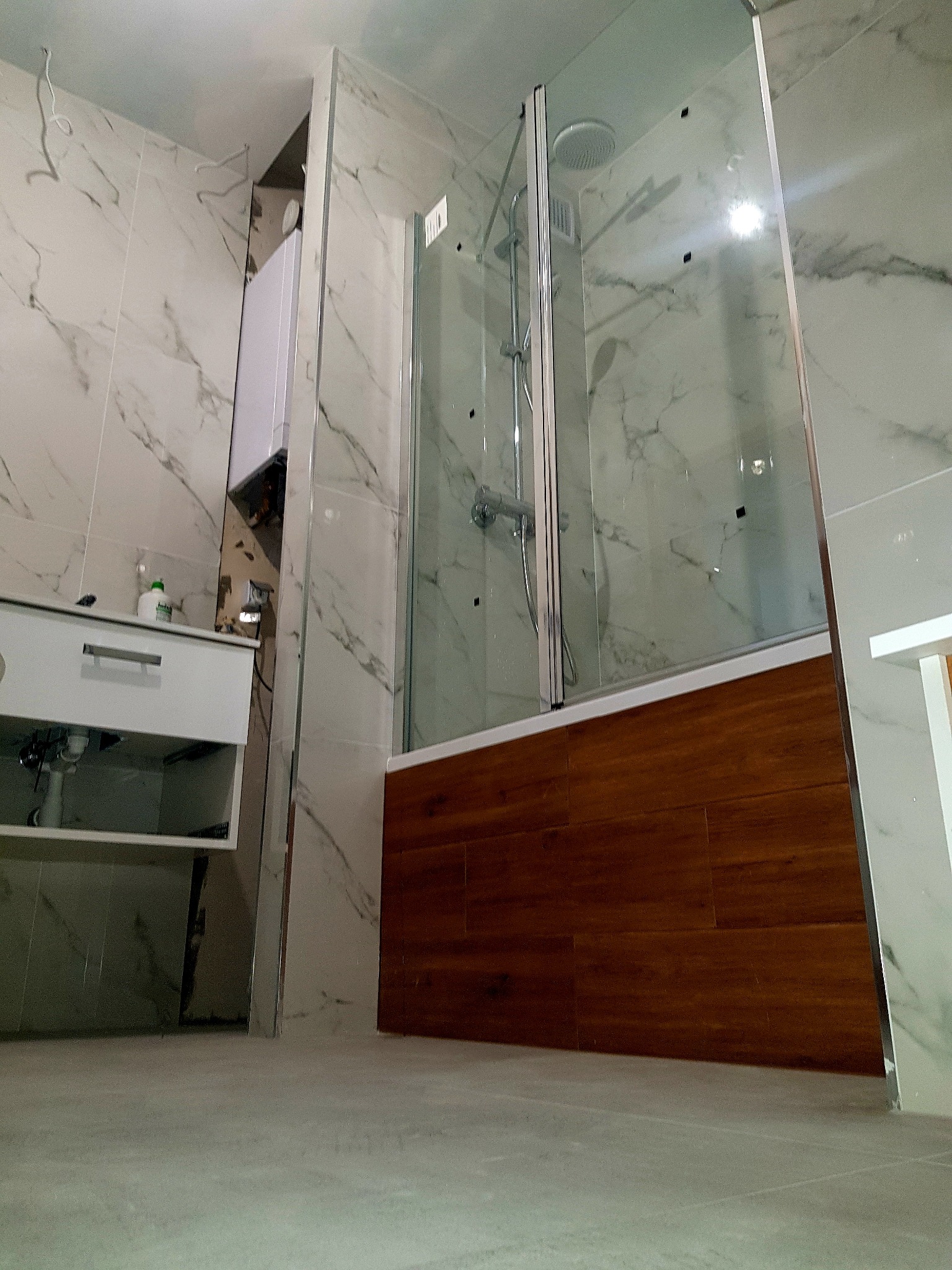 Niedokończona łazienka z marmurowymi płytkami, widać zamontowaną kabinę prysznicową z drewnianym akcentem, białą szafkę pod umywalkę i wystający piec gazowy.