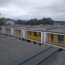 P.D.Tech Płaski Dach PVC Michał Gajda - Nowoczesny budynek z płaskim dachem pokrytym blachą na rąbek stojący, widok z góry na fragment dachu i balkony z żółtymi osłonami. Osoba na balkonie.