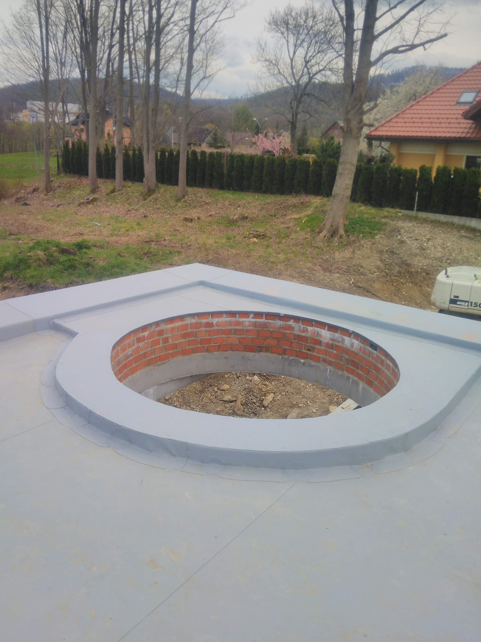 Dach płaski z nietypowym okrągłym otworem wykończonym czerwoną cegłą, zabezpieczony szarą membraną. W tle budynki i drzewa.