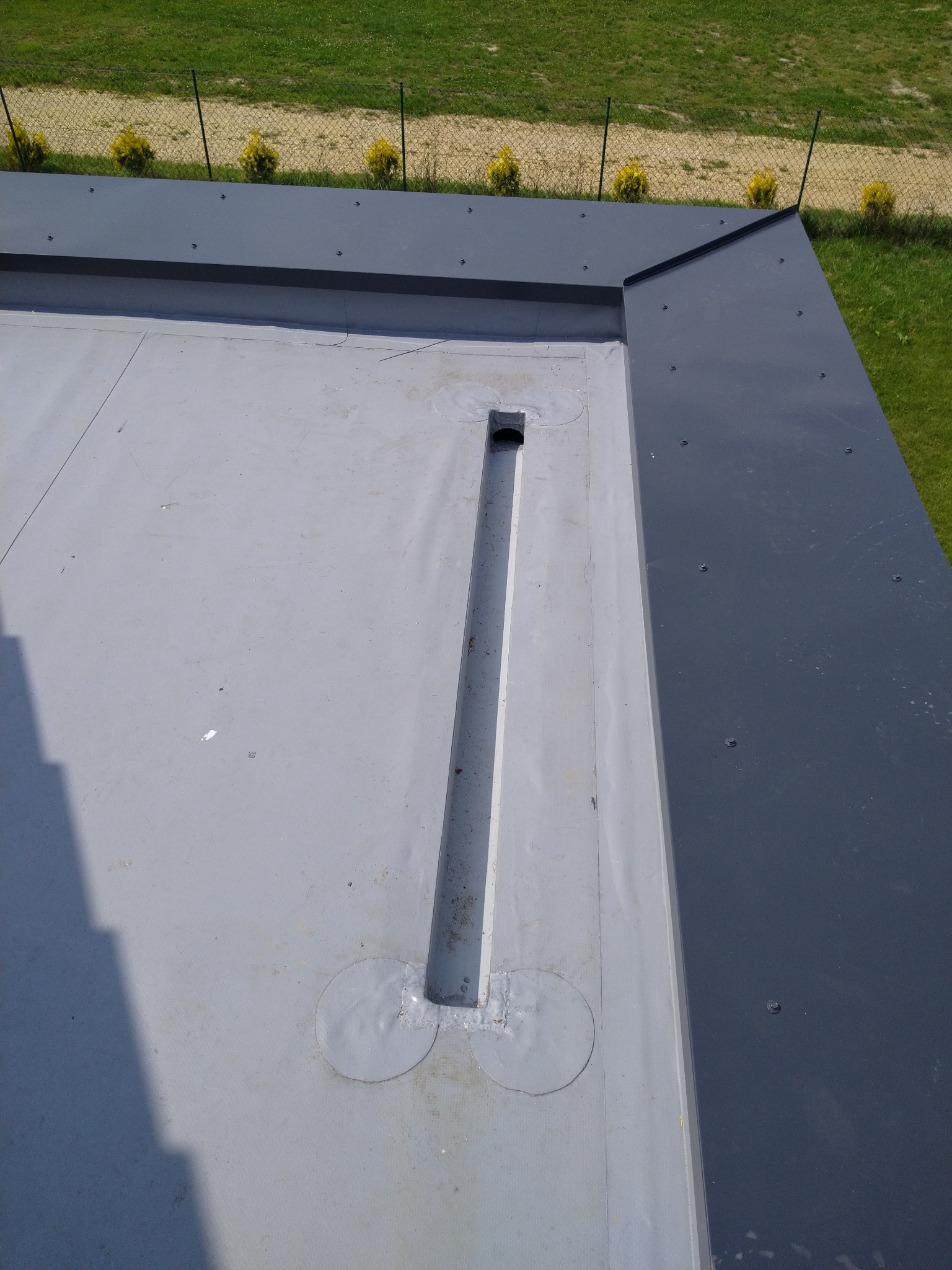 Szary, płaski dach z odpływem liniowym i obróbkami blacharskimi, widok z góry na tle zieleni. Detal wykończenia dachu płaskiego.