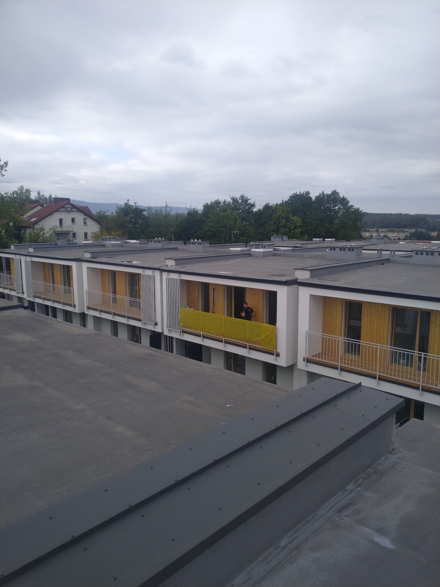 Nowoczesny budynek z płaskim dachem pokrytym blachą na rąbek stojący, widok z góry na fragment dachu i balkony z żółtymi osłonami. Osoba na balkonie.