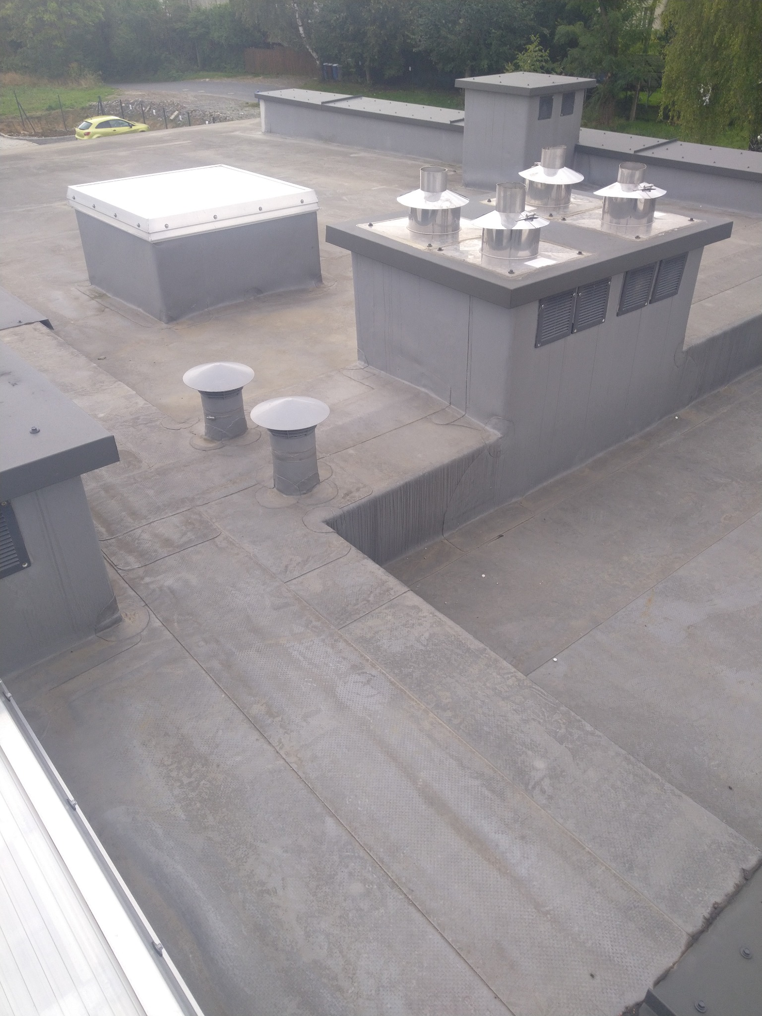 Szary, płaski dach z kominami wentylacyjnymi, świetlikiem i wywietrznikami. Widoczna faktura pokrycia. W tle zieleń drzew i fragment żółtego samochodu.