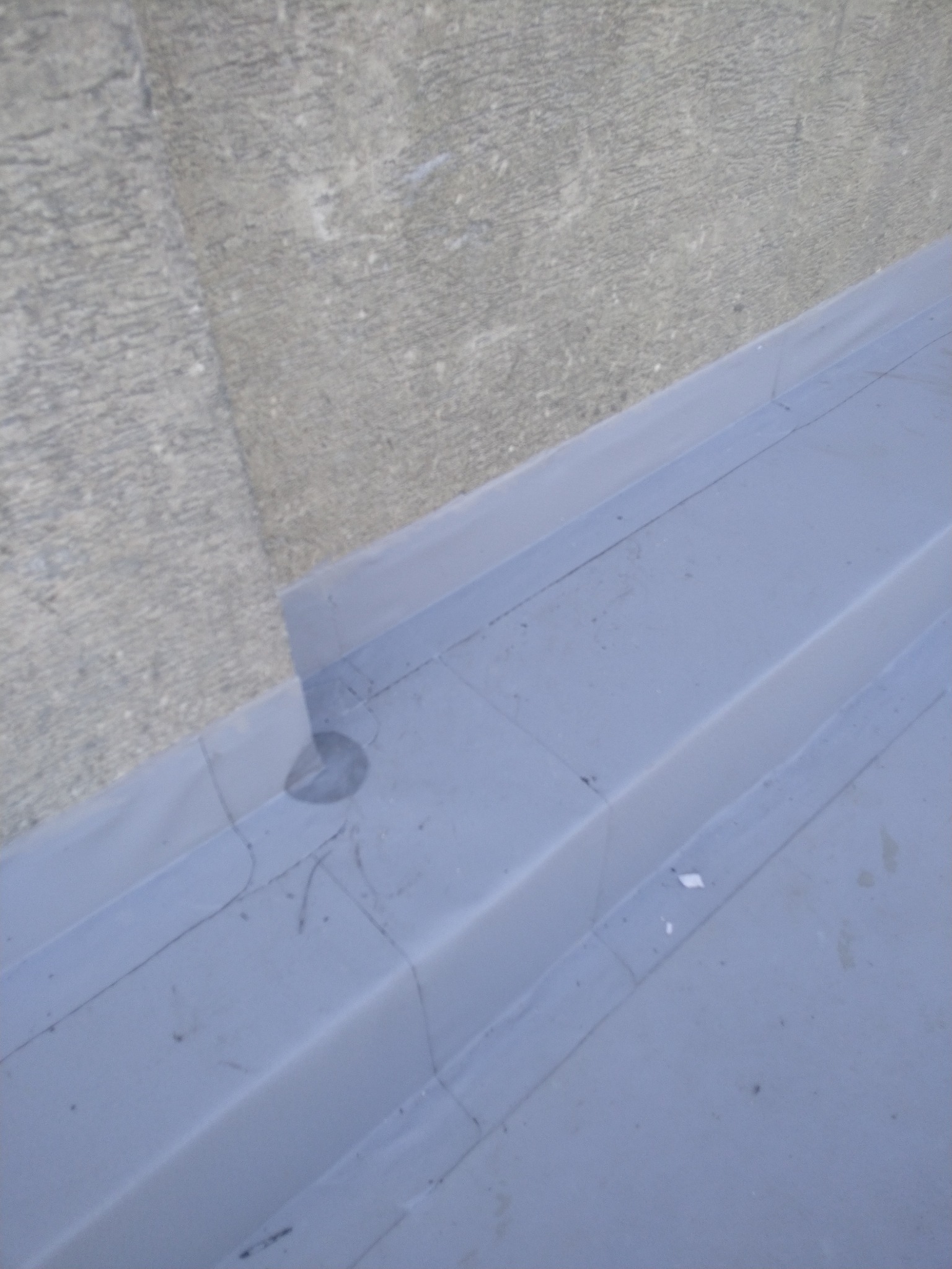 Szary dach kryty papą termozgrzewalną, widok z góry na fragment z widocznymi łączeniami i fakturą papy. Tło stanowi betonowa ściana.