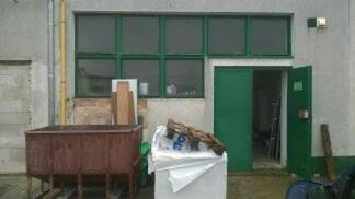 Wejście do warsztatu lub magazynu z otwartymi, zielonymi drzwiami, nad którymi znajduje się duże okno podzielone na kilka mniejszych szyb. Przed wejściem widoczne różne materiały budowlane i pojemnik.