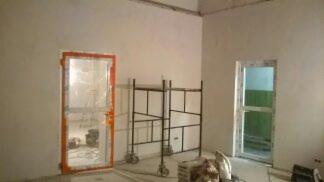 Wnętrze w trakcie prac wykończeniowych: szpachlowane ściany, metalowe rusztowanie, zabezpieczone folią ościeżnice drzwiowe z widocznymi śladami zaprawy, surowy betonowy sufit, podłoga z widocznymi...