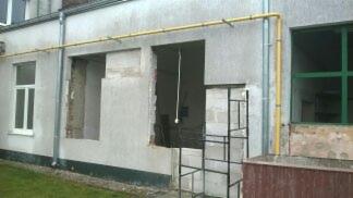 Otwór drzwiowy w trakcie wykuwania w szarej, otynkowanej ścianie budynku. Widoczne fragmenty cegieł i pustaków, żółta instalacja gazowa na elewacji, drabina rusztowania pod otworem.