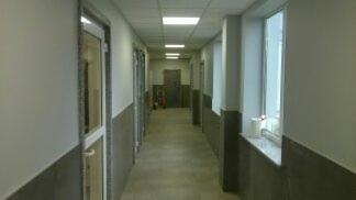 Długi korytarz z szarymi panelami ściennymi do połowy wysokości, białymi ścianami powyżej i białym sufitem z wbudowanym oświetleniem. Po lewej stronie widoczne drzwi z aluminiową ramą, po prawej...