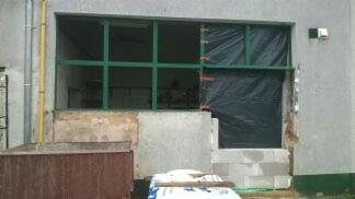 Otwór okienny z zieloną ramą w trakcie montażu, częściowo zakryty czarną folią, widoczne bloczki betonowe i materiały budowlane, ściana zewnętrzna budynku.