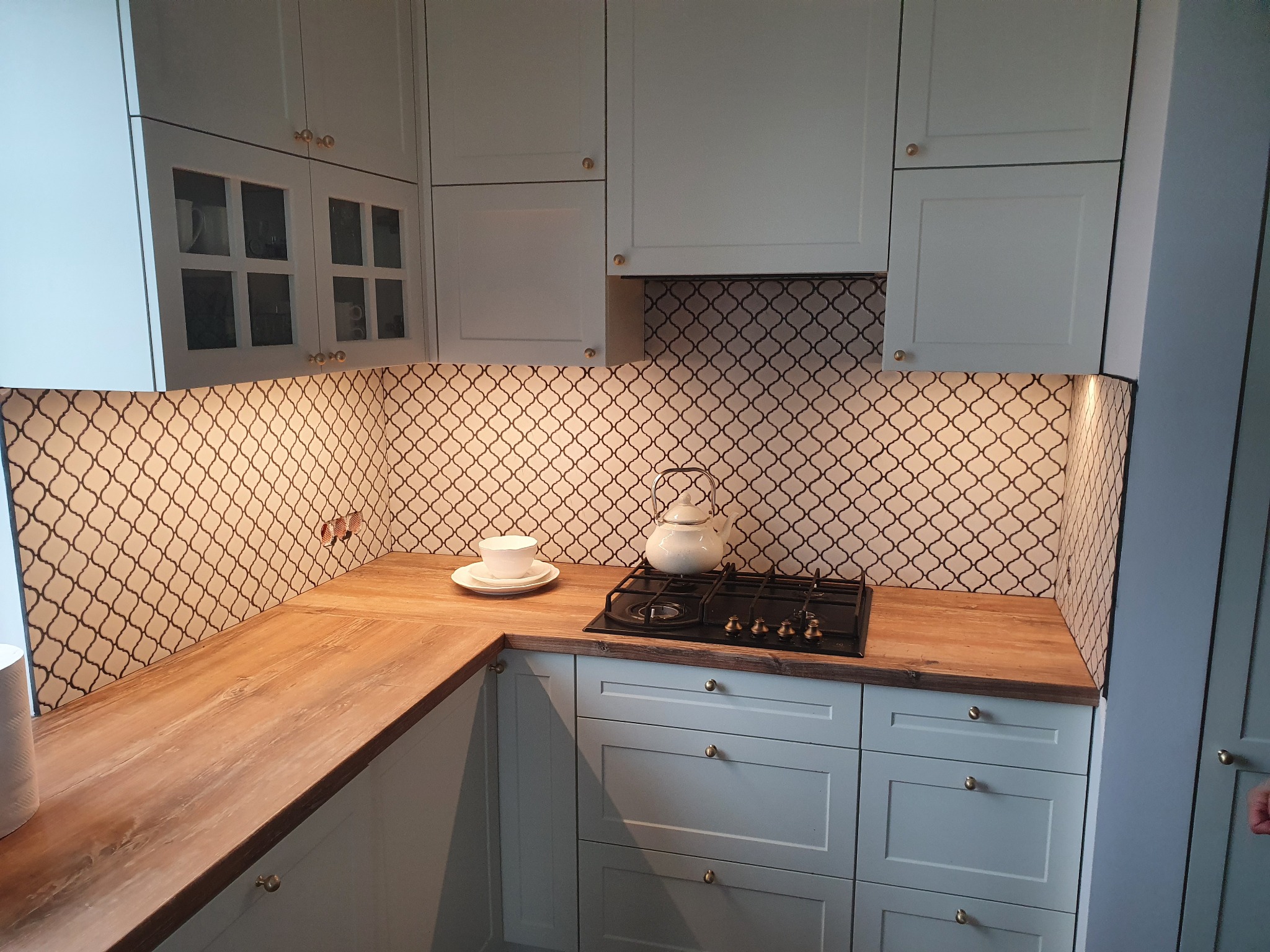 Jasna kuchnia z drewnianym blatem, mozaiką na ścianie, płytą grzewczą i czajnikiem, filiżanka na blacie.