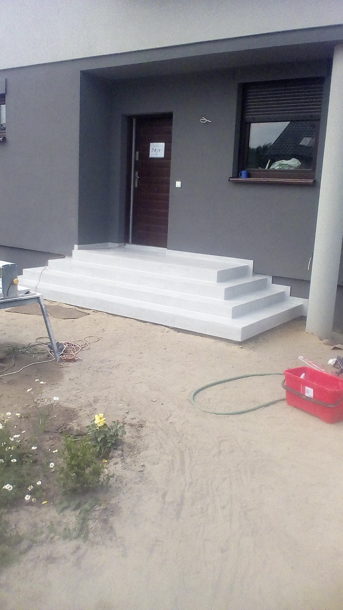 Nowe, białe, wykafelkowane schody wejściowe do domu, w tle szara elewacja i brązowe drzwi z numerem 3A/1, widoczne narzędzia i wiadro.