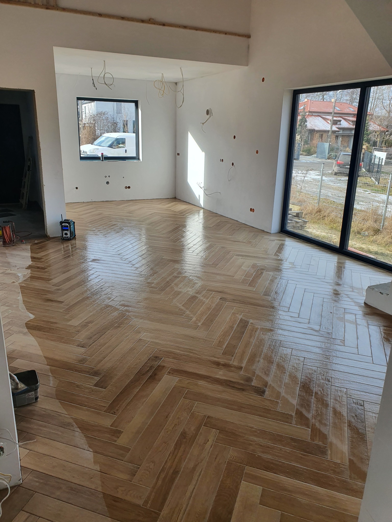 Błyszcząca podłoga z płytek ułożonych w jodełkę w jasnym, przestronnym wnętrzu w trakcie wykańczania, widoczne przewody elektryczne wystające ze ścian i okno z widokiem na zaparkowany samochód.