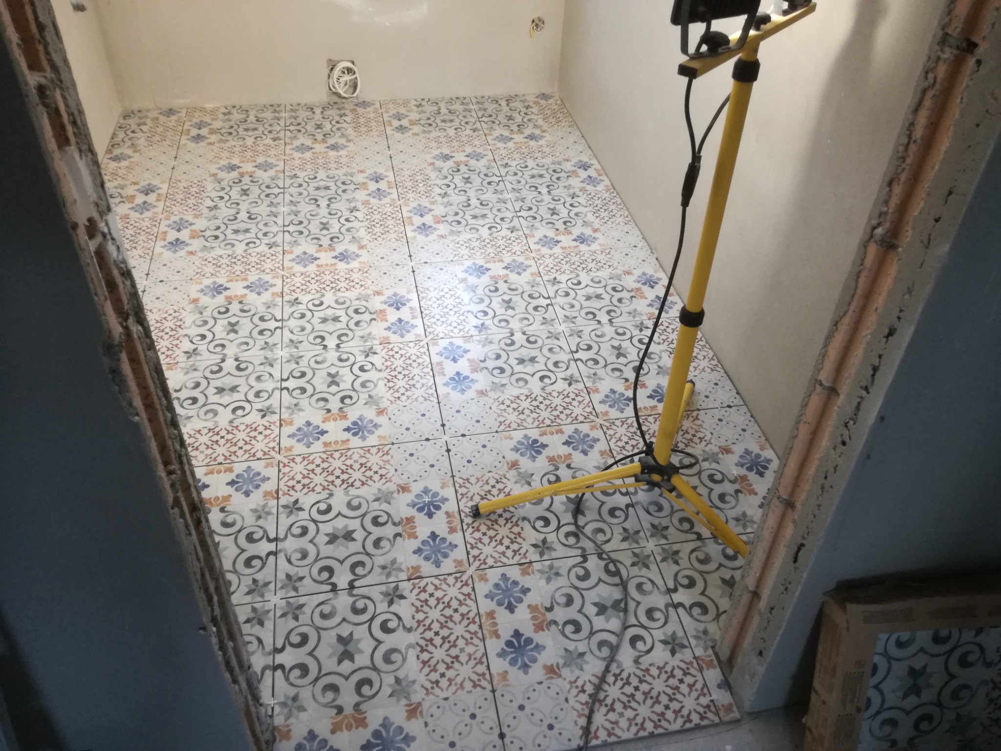 Ułożona podłoga z wzorzystych płytek w łazience, widoczna żółta lampa budowlana i surowy ościeżnica drzwi, instalacja elektryczna w ścianie.