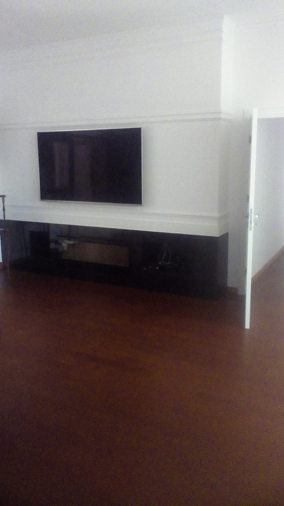 Salon z ciemną, lakierowaną podłogą, białe ściany z listwami dekoracyjnymi, wiszący telewizor nad czarną, długą szafką RTV, otwarte białe drzwi.