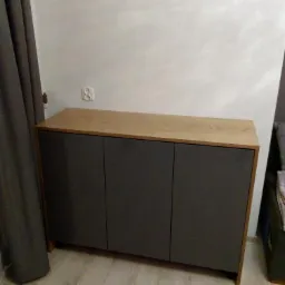 Nowoczesna komoda z trzema ciemnoszarymi frontami i blatem w kolorze jasnego drewna, stojąca przy ścianie z widocznym gniazdkiem elektrycznym i szarą zasłoną.