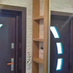 Wąski, drewniany regał z półkami w zabudowie szafy przesuwnej, obok ciemnych drzwi wejściowych w Więciórce, na tle ściany z dekoracyjną okładziną kamienną.