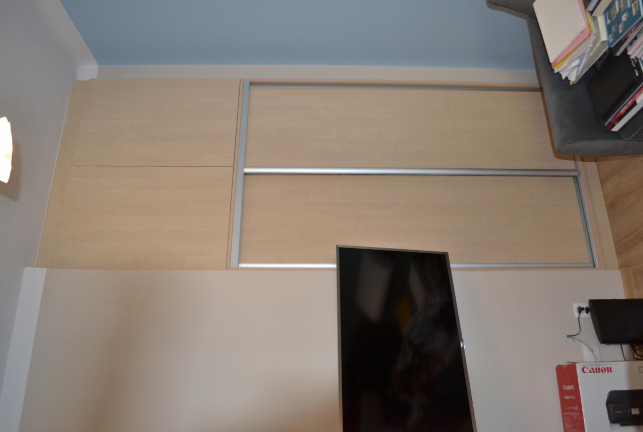 Jasna szafa przesuwna z drewnopodobnymi frontami, wbudowana od podłogi do sufitu, z widocznym telewizorem na ścianie i fragmentem szarego fotela z książkami.