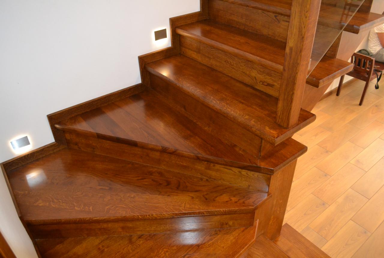 Polerowane, dębowe schody z podświetleniem LED w ścianie, widok z góry na stopień zabiegowy, parkiet drewniany w tle.