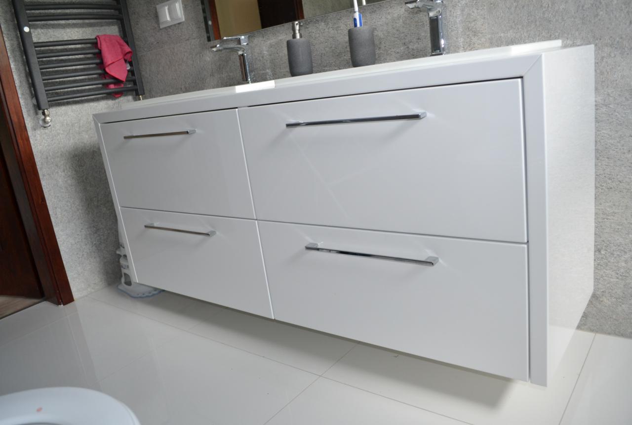 Biała szafka pod umywalkę z czterema szufladami i chromowanymi uchwytami, widoczna z bliskiej perspektywy, z elementami instalacji wodnej i grzejnikiem drabinkowym w tle.