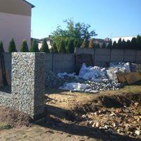 Słup z gabionów w ogrodzie, widoczne przygotowane kamienie do wypełnienia, w tle ogrodzenie z prefabrykatów betonowych i zabudowa jednorodzinna w oddali.