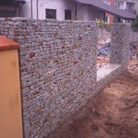 Mur oporowy z kamienia w metalowej siatce gabionowej, z widocznym fragmentem słupka ogrodzeniowego w tle i nierównym terenem wokół konstrukcji.