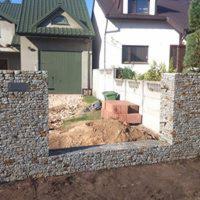 Fragment ogrodzenia gabionowego z szarego kamienia na tle domu z zielonym dachem i garażem, widoczne przygotowane podłoże i materiały budowlane.